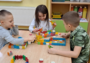 Dwóch chłopców i dziewczynka bawią się figurkami dinozaurów i zbudowanymi z klocków budowlami.